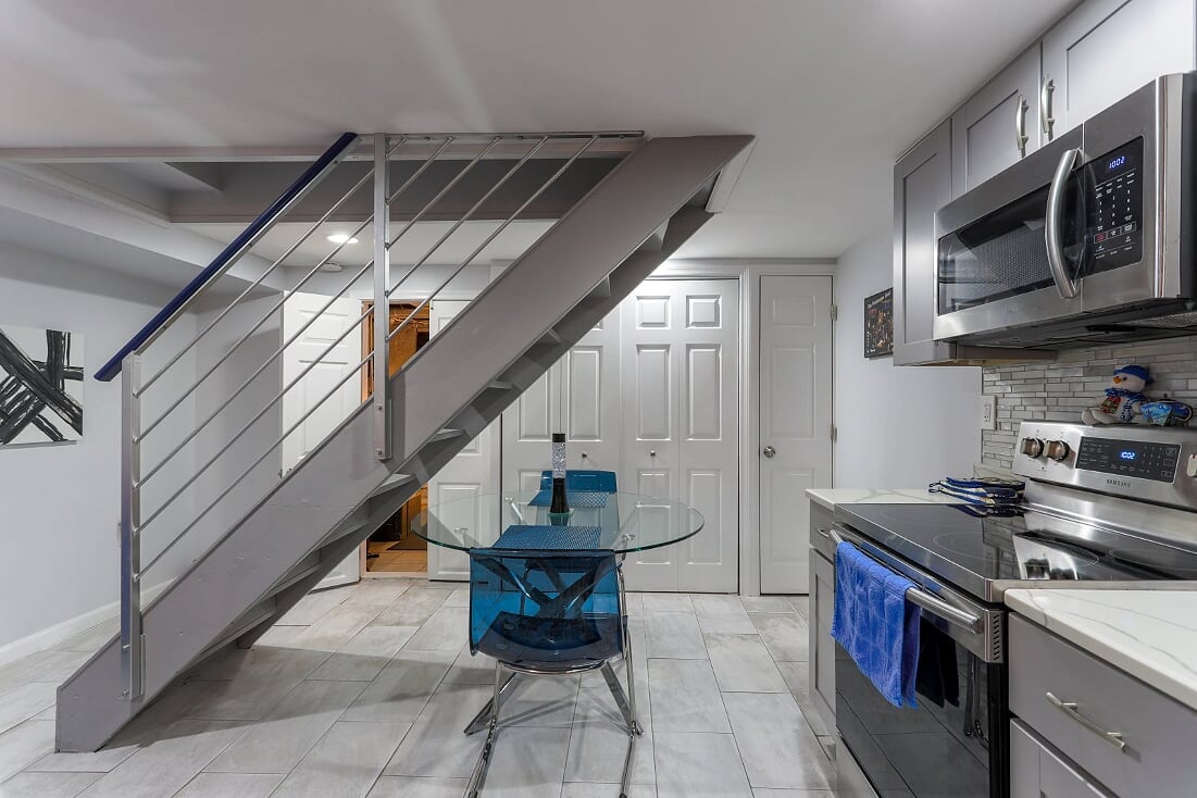 The kitchenette area features a glass dining table surrounded by blue chairs, positioned below a staircase. Stainless steel appliances, including an oven and microwave, are visible along the wall, complemented by a small decorative piece. A door leads to another room, enhancing accessibility.