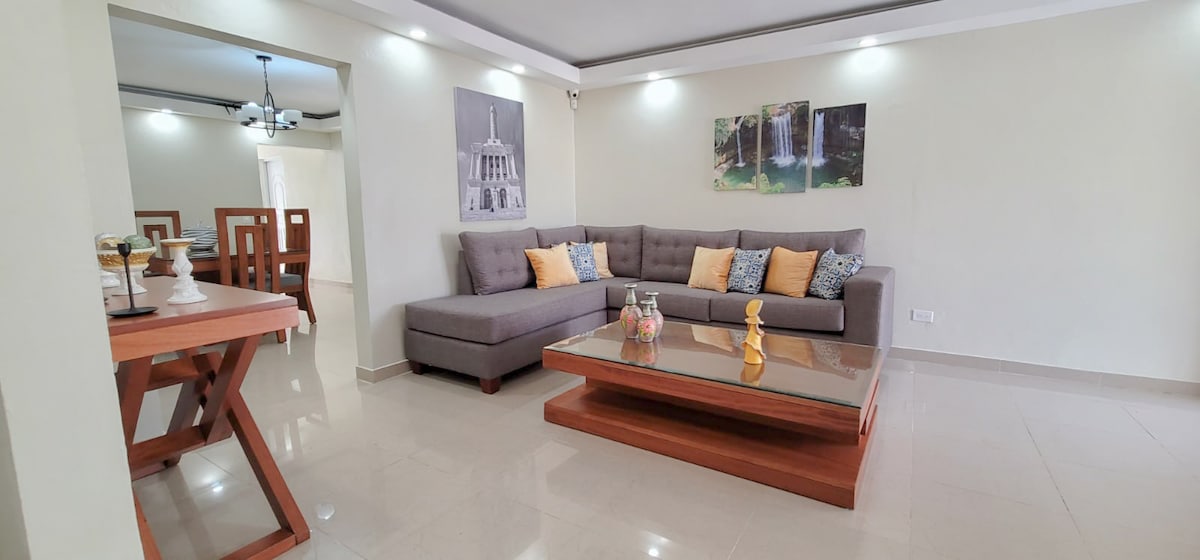 A spacious living room is presented, featuring a large grey sectional sofa adorned with yellow and patterned cushions. A wooden coffee table sits in the center, complemented by decorative items. Light is softly illuminated by ceiling fixtures, highlighting the neutral walls and tiled flooring.