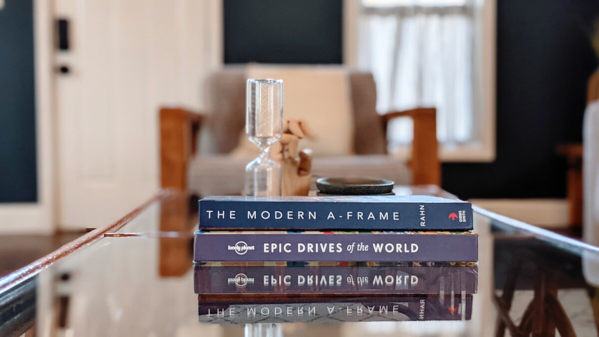 A coffee table is featured in a well-lit living room, displaying a stack of travel and design books. A glass hourglass is positioned atop the books, with the blurred background revealing a cozy seating area and dark blue walls.