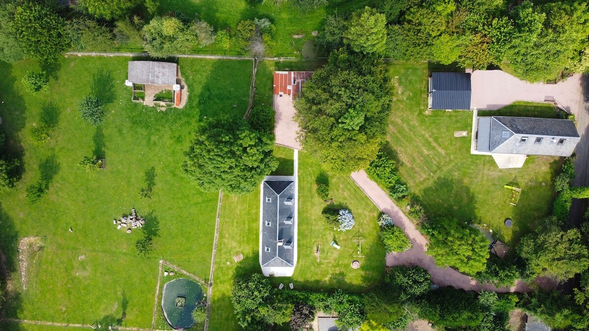 An aerial view captures the spacious grounds of the property, featuring well-maintained green lawns, a pond, and multiple structures including a main building and outbuildings. Pathways meander through the landscape, bordered by trees and greenery.