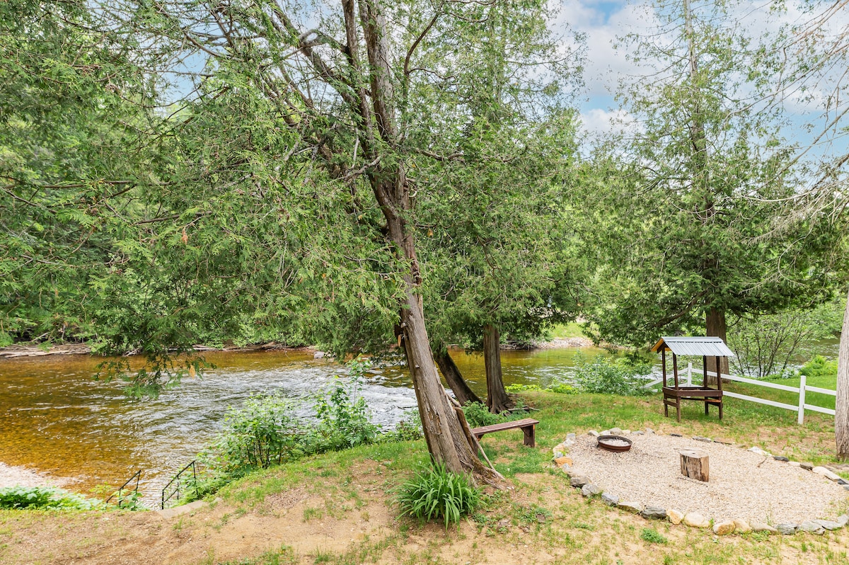 A serene outdoor area features a fire pit surrounded by stones, ideal for gatherings. Nearby, a small shelter and seating are positioned under tall trees, providing shade. The calm river flows gently in the background, complemented by lush greenery along the banks.