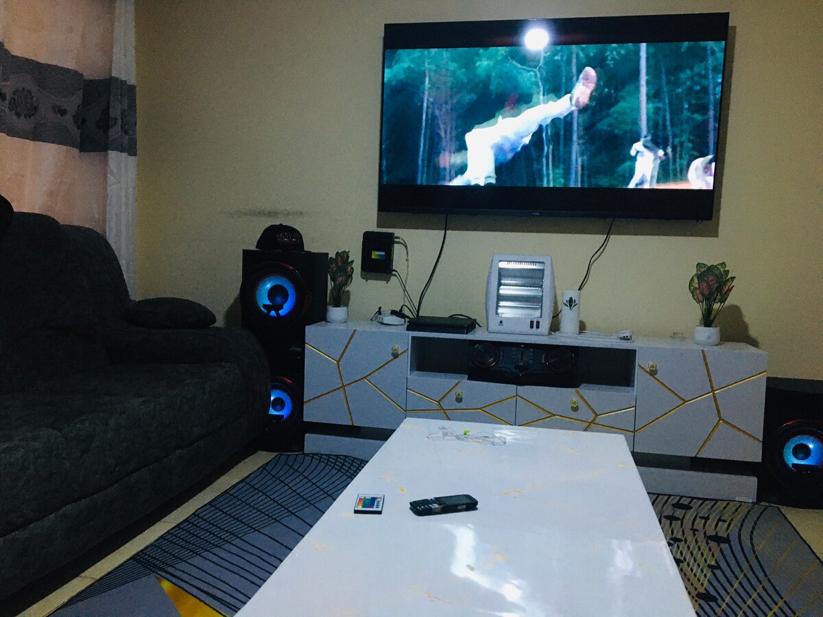 A modern living area features a large television mounted on the wall, displaying a scene from a film. A dark sofa is positioned to the side, with a sleek coffee table in front. Audio speakers are placed beside the furniture, enhancing entertainment options.