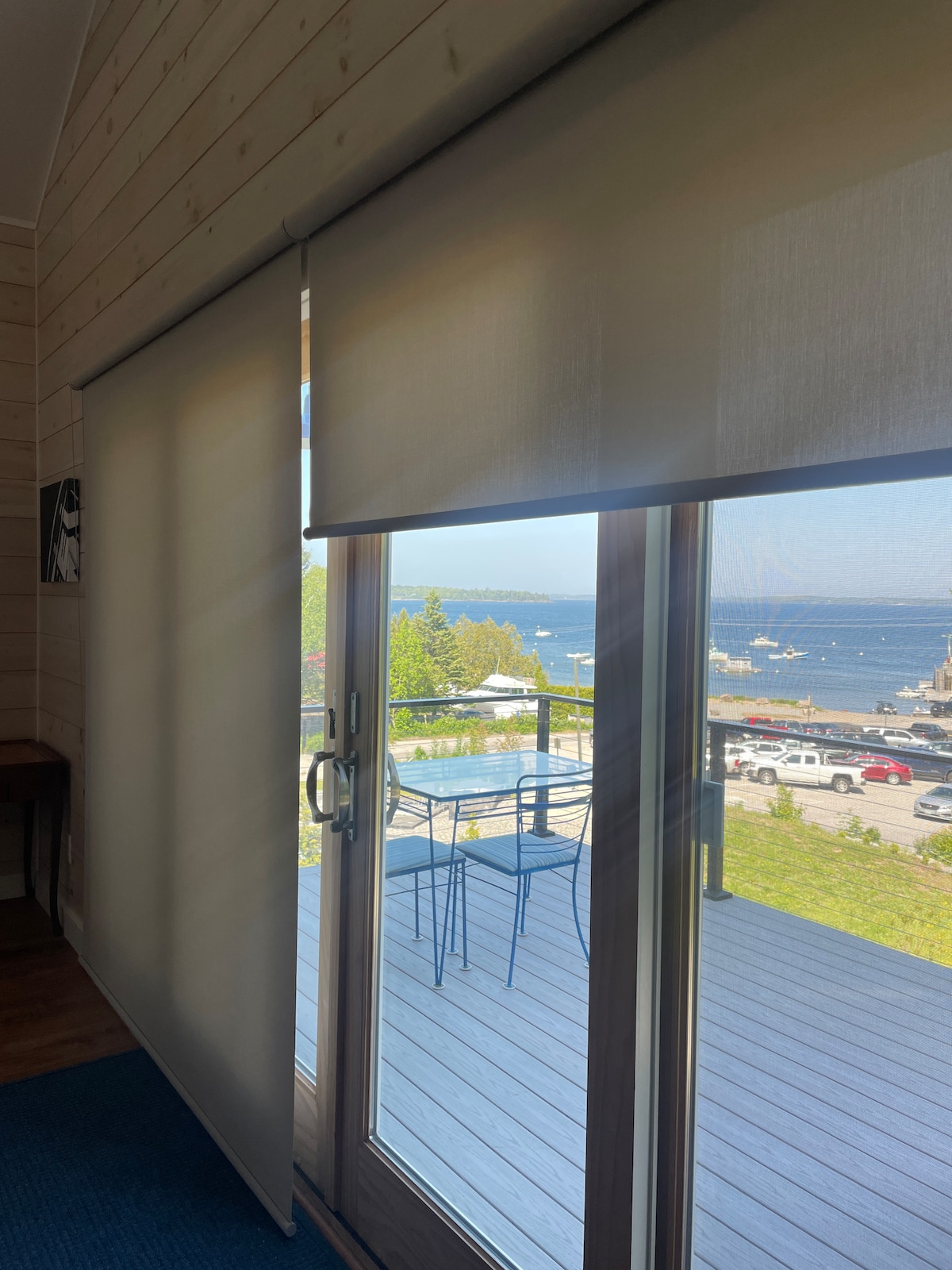Sliding glass doors are revealed with blinds drawn, showcasing a view of the outdoor deck and scenic waterfront. Blue chairs and a small table are visible on the deck, overlooking the calm waters where boats are docked.