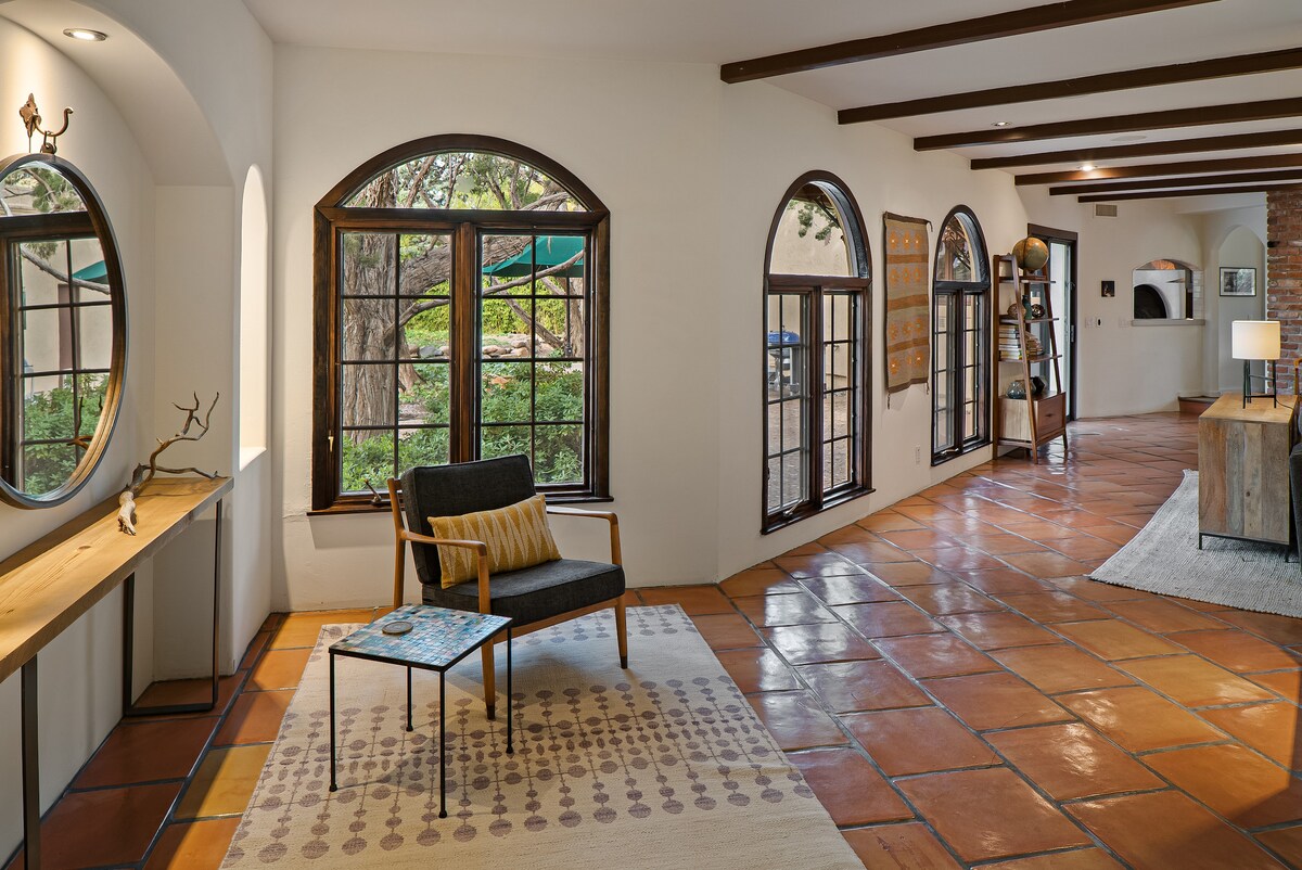 A spacious living area features arched windows that frame views of the landscaped surroundings. A modern armchair is paired with a small side table on a patterned rug over terracotta tiles, enhancing the inviting aesthetic of the hacienda-style home.