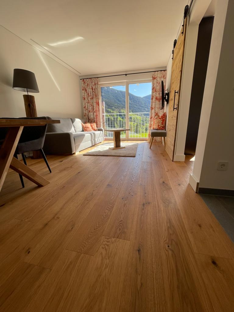 A spacious living area is visible, featuring a sofa and a small table set next to a large window that frames a mountain view. Natural light highlights the wooden flooring and the warm tones of the furnishings, while floral curtains add a touch of color.