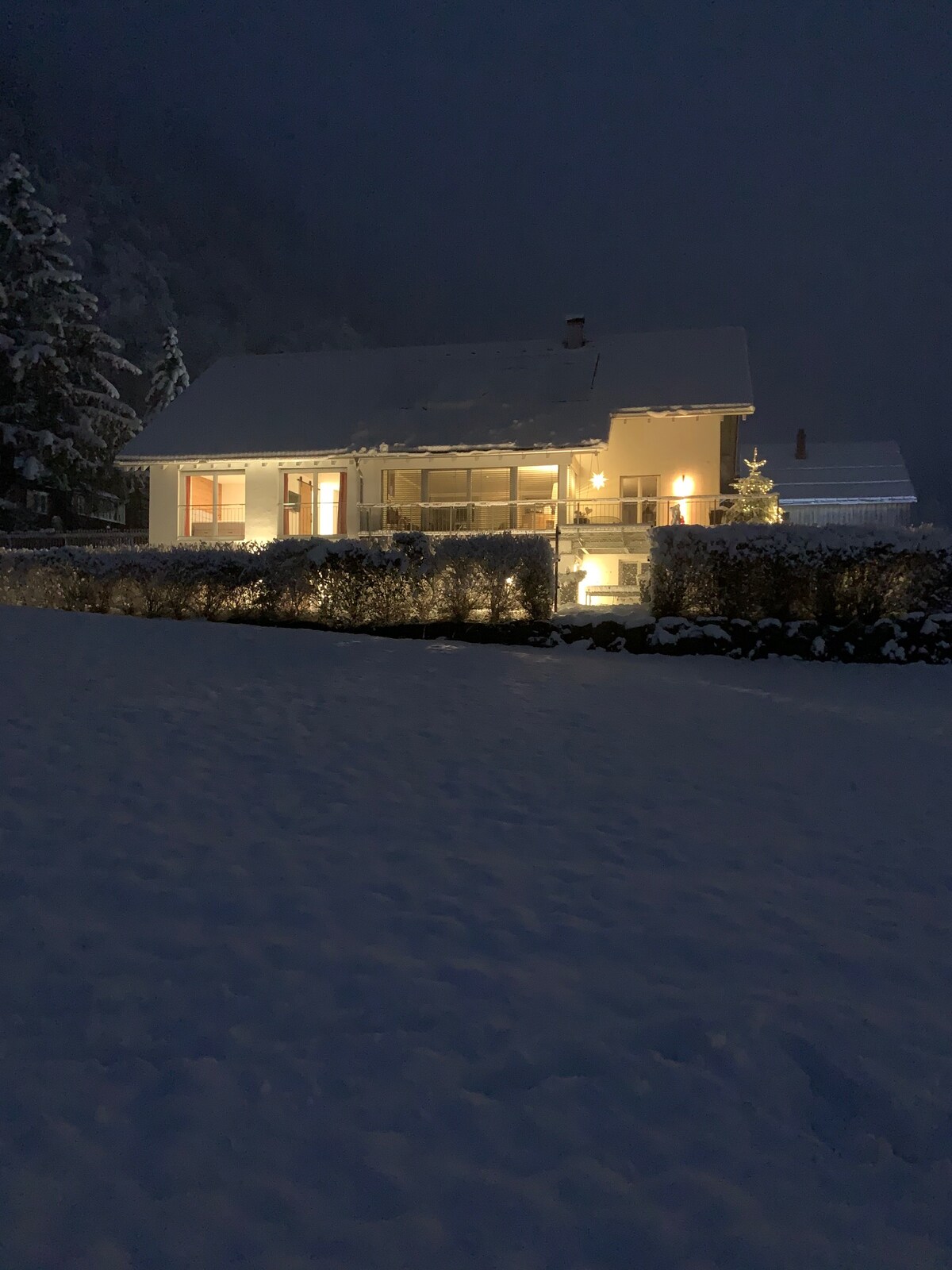 A charming house is illuminated against the winter night, with soft lighting highlighting its exterior. The surrounding snow-covered landscape enhances the serene ambiance. A row of shrubs outlines the property, creating a sense of privacy while snow blankets the ground.