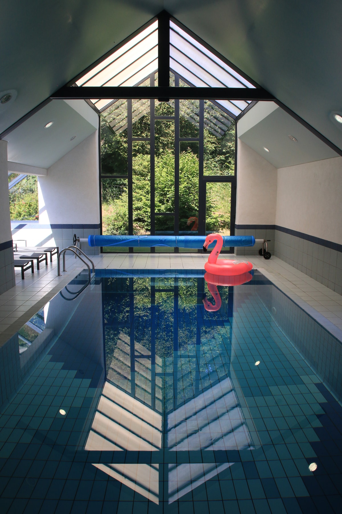 An indoor heated pool is surrounded by large glass windows, offering views of the greenery outside. A pink inflatable flamingo floats in the clear water, while steps lead into the pool area. The clean tiled space features a blue slide and ample natural light.