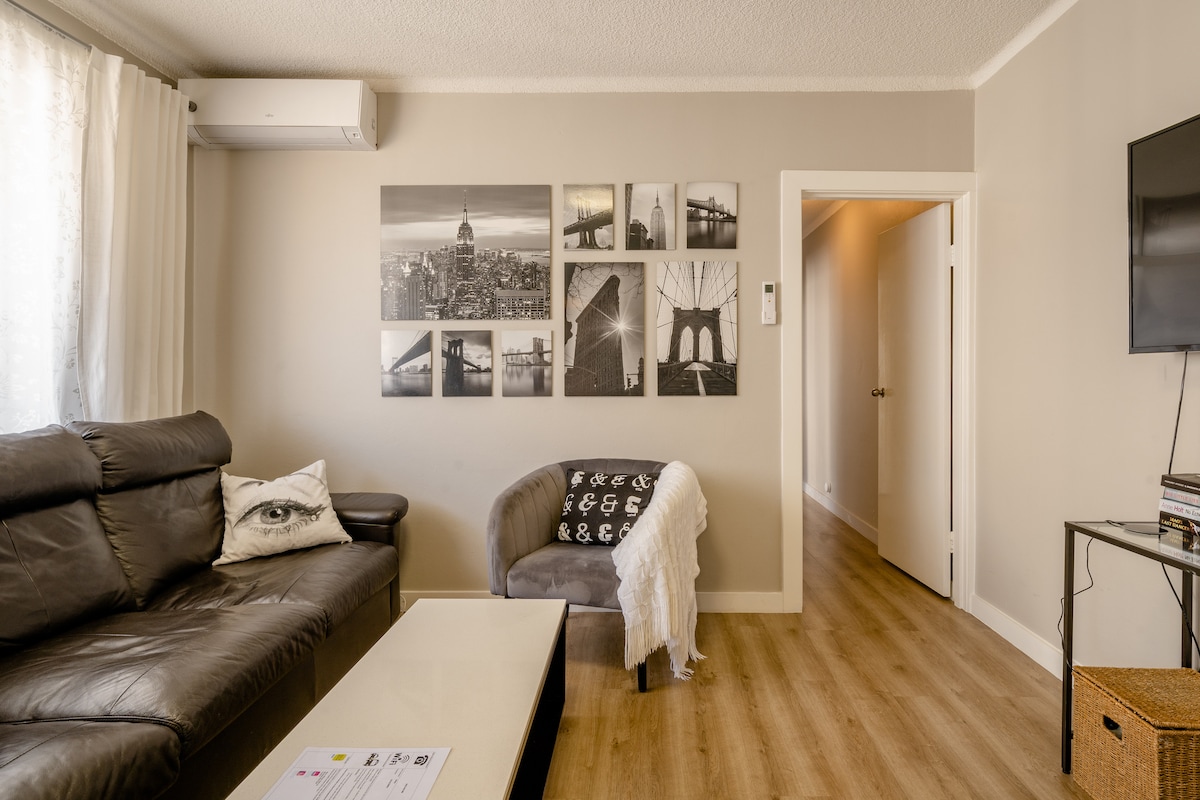 A comfortable living area features a dark leather sofa and a cozy accent chair. A series of black-and-white photographs adorns the wall, showcasing various cityscapes. Natural light filters through sheer curtains, and a small coffee table sits in front, completing the inviting atmosphere.