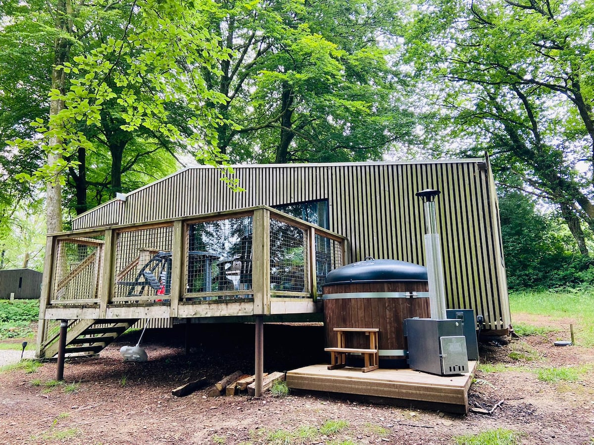 A modern wooden lodge is set within a forested area, featuring a spacious deck with steps leading up to the entrance. A private Nordic bath is placed on the ground beside the lodge, surrounded by greenery. Large windows allow natural light to illuminate the interior.
