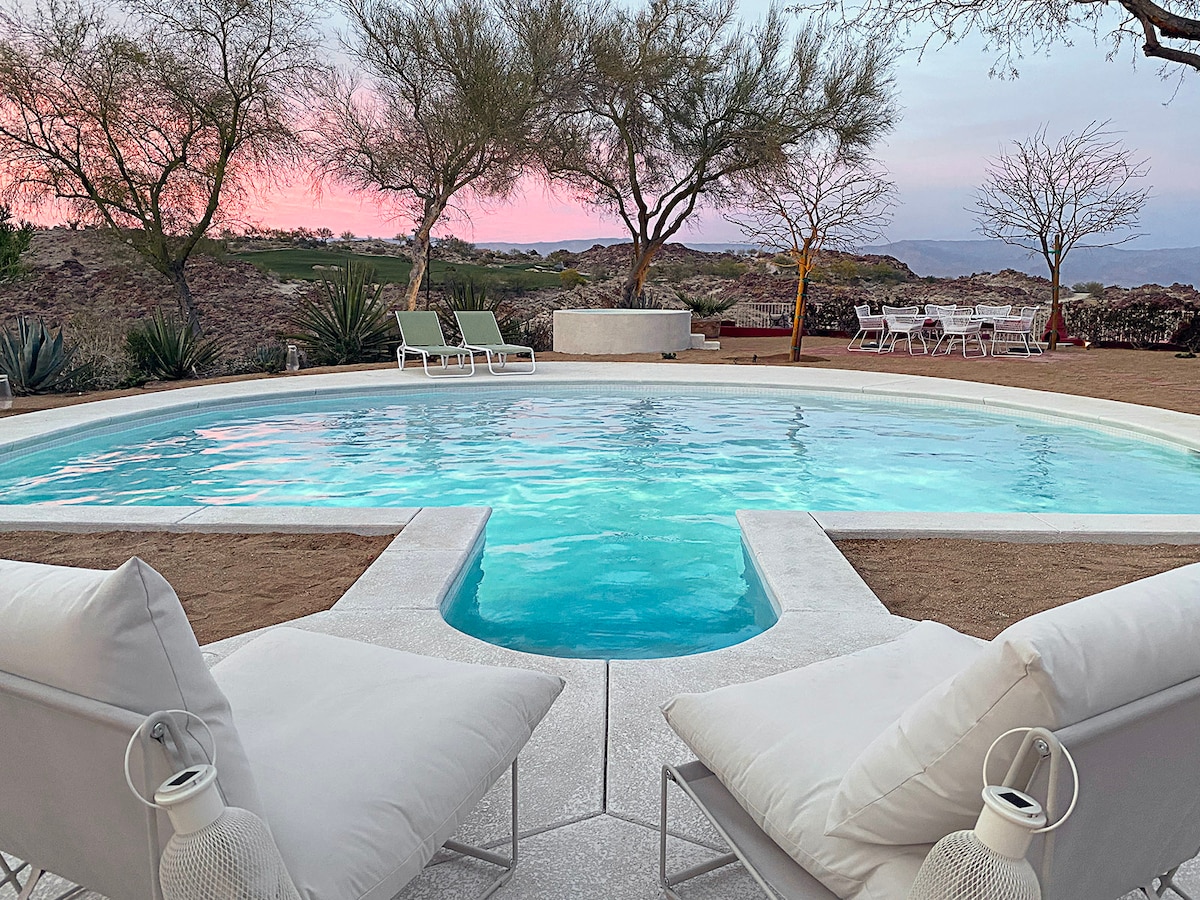 A serene pool area is framed by soft seating and chaise lounges, inviting relaxation. The water reflects soft shades of blue, while the backdrop features desert trees against a pastel sky. A dining setup can be seen in the distance, enhancing the peaceful ambiance.