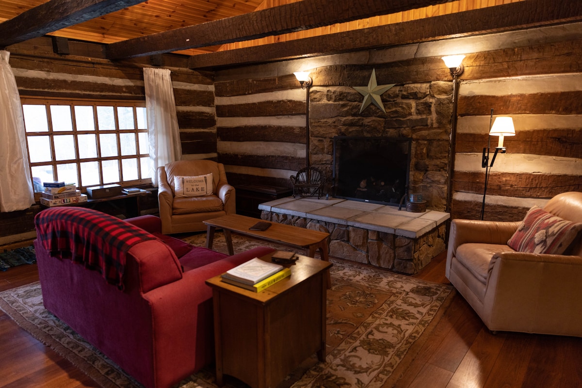 A cozy living area features wooden beams and rustic log walls, creating a warm ambiance. Two seating areas are present: a crimson sofa with a plaid throw and a light-toned armchair. A stone fireplace, accented by a decorative star, serves as a focal point, surrounded by soft lighting.
