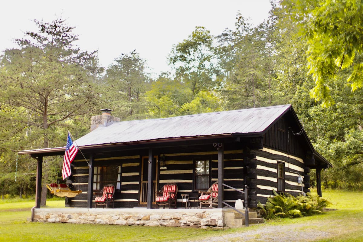 Small cabin exterior of this West Virginia lake house rental with chairs and swing on the porch, nestled in the woods.