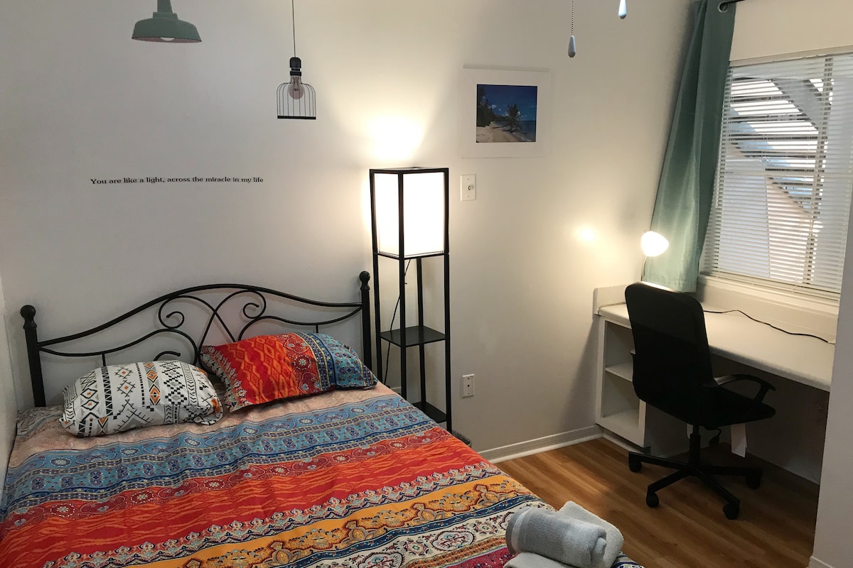 A private room is furnished with a full-sized bed adorned with vibrant bedding. A desk with a chair is positioned next to a window displaying natural light, complemented by floor and overhead lamps. Decorative wall art adds a subtle character to the space.