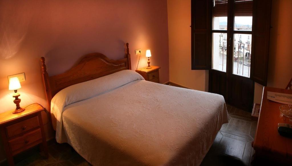 A comfortable bedroom features a large bed with a white quilt and wooden furnishings. Two lamps provide soft lighting on either side of the bed, and a window offers a view of the outside, framed by wooden shutters.