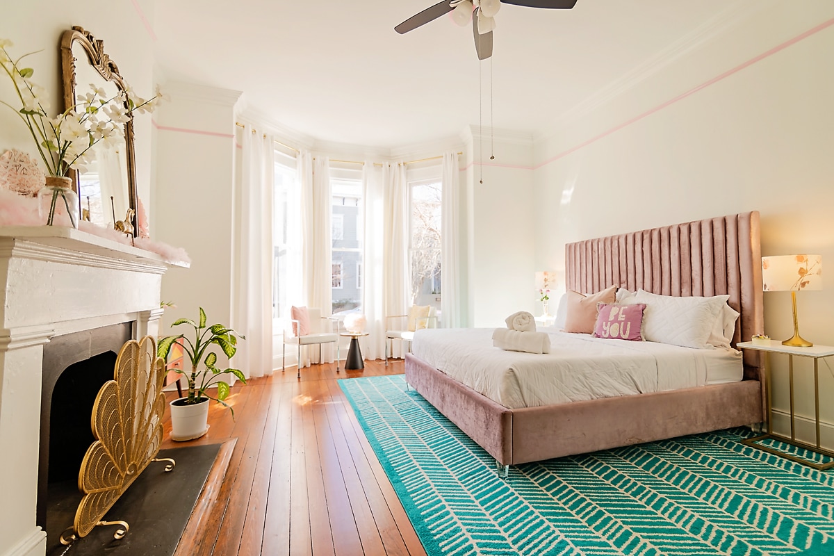 A bright bedroom features a plush king bed with a pink velvet headboard. Large windows allow natural light to fill the space, illuminating the elegant décor. A cozy area with comfortable seating is situated near the windows, complemented by a charming fireplace and decorative plants.