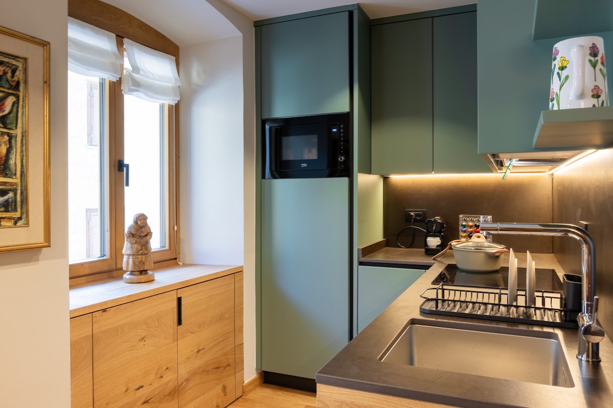 A modern kitchen area is showcased, featuring green cabinetry and sleek countertops. A built-in microwave is mounted above the counter, and a window allows natural light to brighten the space. A dish rack with dishes sits next to a stainless steel sink, enhancing functionality.