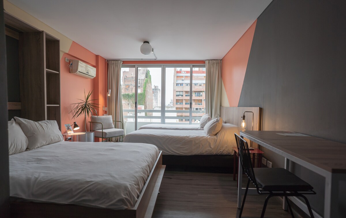 A modern room features two spacious beds, soft white linens, and neutral-toned décor. A wooden desk with a black chair sits beside a large window, allowing natural light to fill the space. A small indoor plant adds a touch of greenery.
