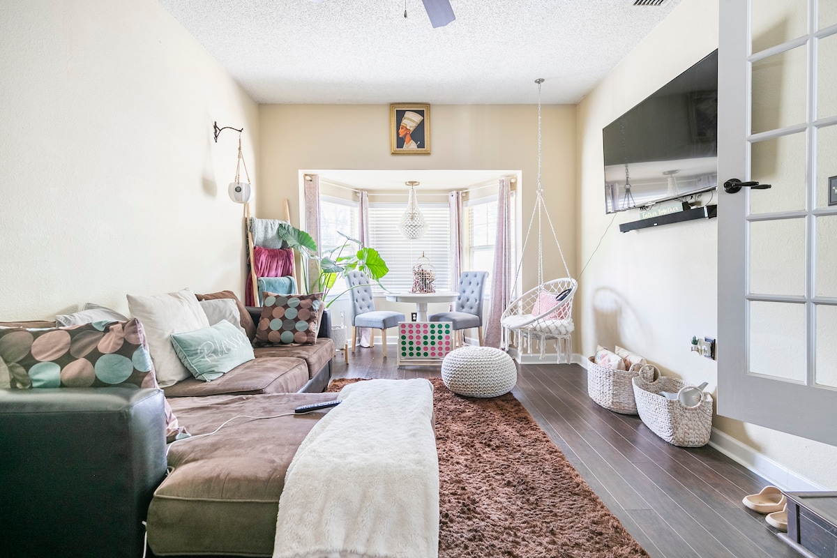 A cozy living area features a plush brown sectional sofa adorned with decorative pillows. A light area rug complements dark flooring. Large windows allow natural light, while indoor plants add a touch of greenery. A wall-mounted TV and colorful elements enhance the inviting ambiance.