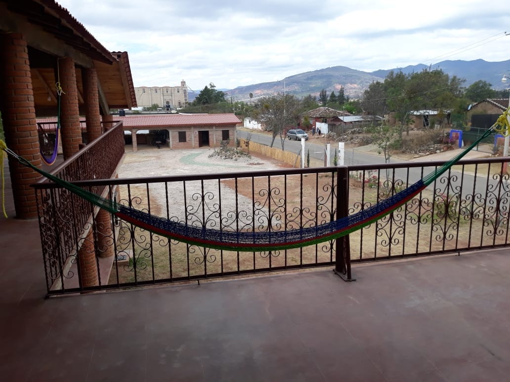 A balcony is shown featuring an intricately designed railing, with a colorful hammock suspended across it. Below, an expansive outdoor space is visible, including a gravel area and distant buildings set against a backdrop of rolling hills under a cloudy sky.
