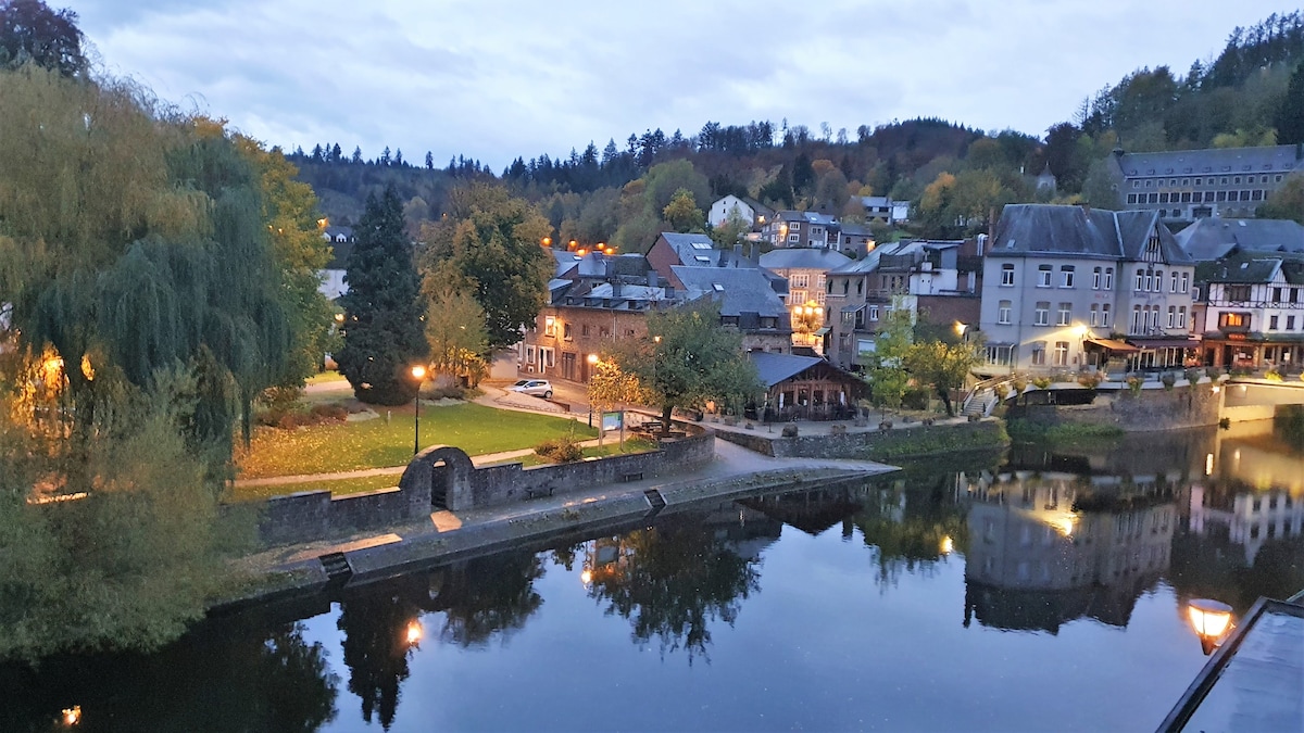 A scenic view of the town along the river Ourthe during twilight. Soft light reflects off the water, highlighting the surrounding trees and buildings. Grassy areas and pathways are visible, with a sense of tranquility enhanced by the soft illumination from street lamps.