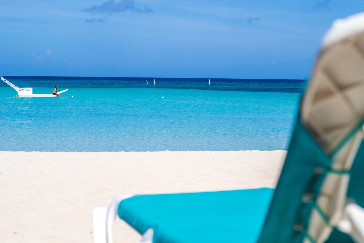 A serene beach scene featuring soft white sand and turquoise waters stretching into the horizon. A nearby beach chair is positioned to overlook the calm sea, where a person is seen engaging in water activities, enhancing the inviting coastal ambiance.