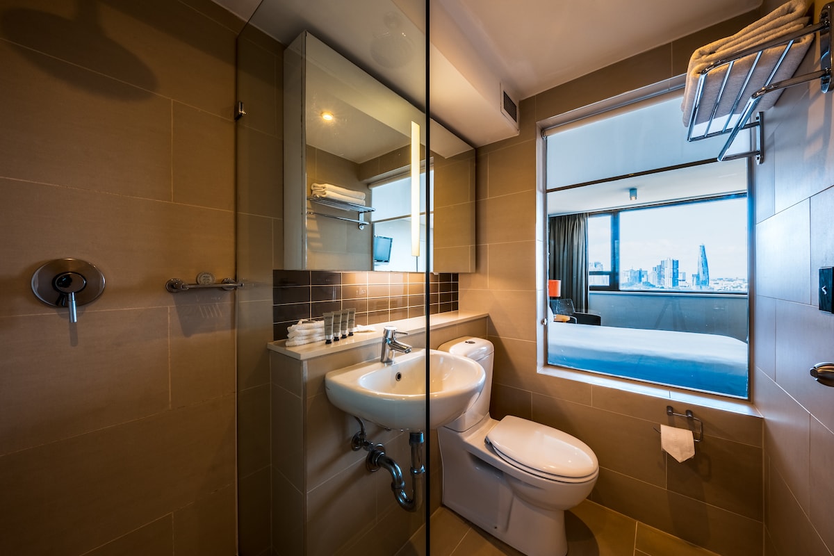 A modern ensuite bathroom features a spacious vanity with a large mirror, complemented by a contemporary toilet and shower area. The window offers a view into the room, allowing natural light to enhance the clean, sleek lines of the design.