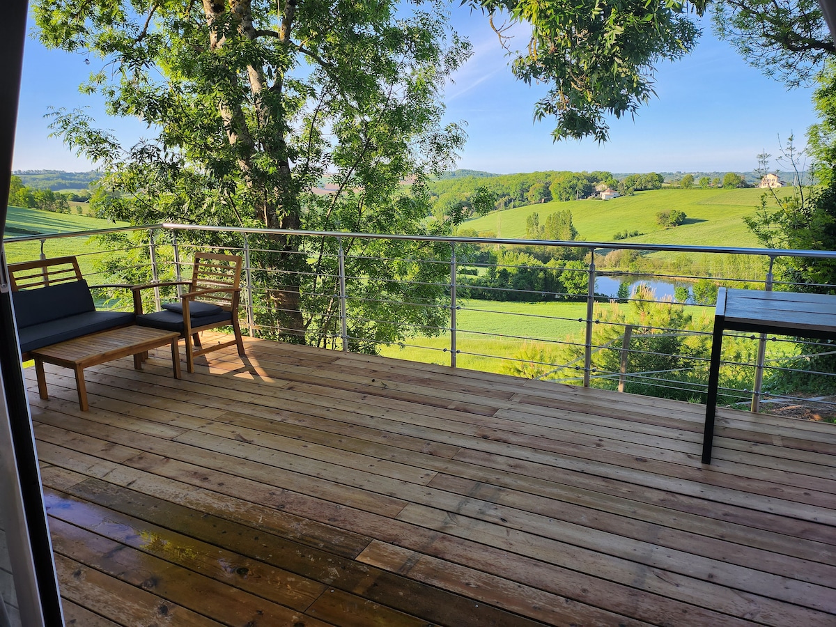 A wooden terrace features two chairs and a table, surrounded by lush greenery. A scenic view of rolling hills and a pond is visible beyond the railing, with sunlight illuminating the natural landscape.
