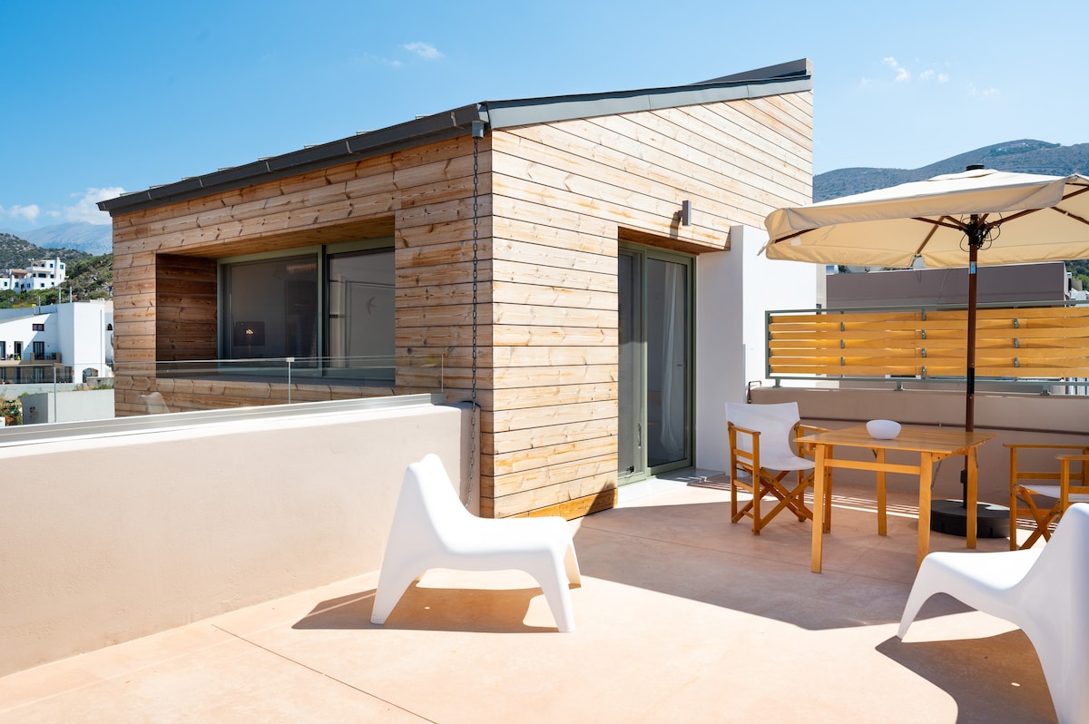 A modern building features a wooden exterior with large windows offering natural light. A spacious terrace is equipped with a dining table and chairs, along with a beige umbrella for shade, providing an inviting area for outdoor relaxation.