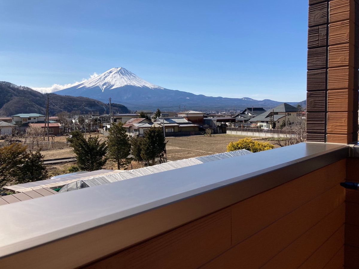A serene view is presented from the balcony, showcasing Mount Fuji in the background. The landscape includes distant houses, trees, and open fields under a clear blue sky. The balcony railing is visible, adding to the overall sense of outdoor space.