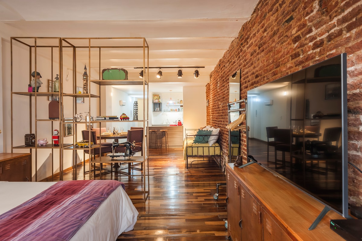 An open living space features exposed brick walls and wooden floors. A spacious bed is positioned beside a display shelving unit. A dining area with stylish chairs and a modern kitchen can be seen in the background, complemented by a large television mounted on a wooden console.