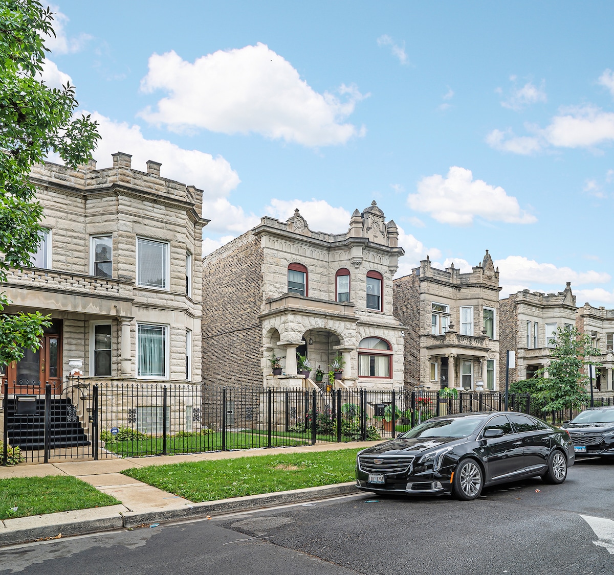A row of historical Greystone buildings is presented, showcasing intricate architectural details and decorative stonework. Well-maintained front lawns feature greenery and plants. A parked car is visible on the street, and a clear blue sky adds to the scene.