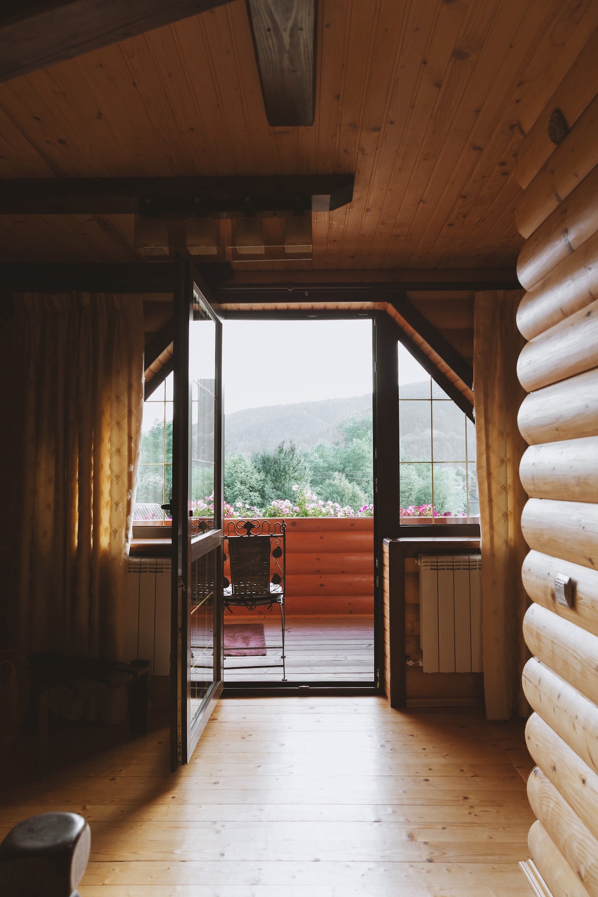 A wooden interior features a doorway leading to a balcony, framed by large windows. Sunlight enters the space, illuminating the wooden walls and floor, while greenery and flowers are visible outside. Soft curtains provide a gentle touch, enhancing the natural setting.