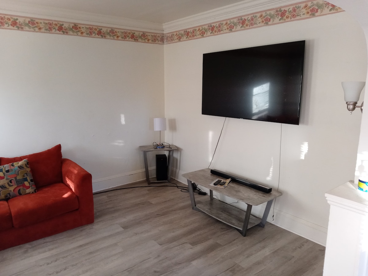 A cozy living area is depicted with an orange sofa placed beside a small side table and lamp. A large flat-screen television is mounted on the wall opposite a simplistic coffee table, enhancing the space's functionality. Light enters through a nearby window, adding brightness to the room.