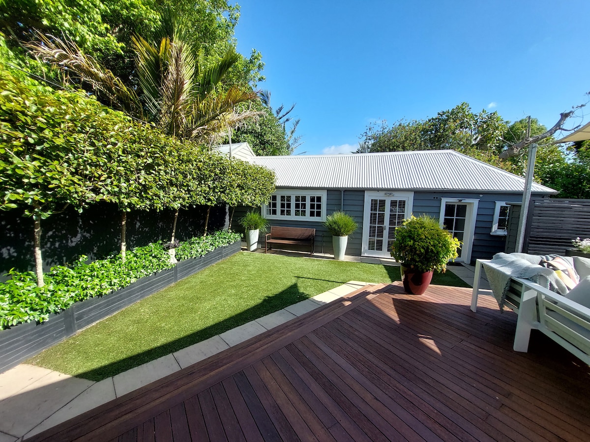A garden studio is visible beyond a manicured lawn, surrounded by vibrant greenery and palm trees. The wooden deck foreground features outdoor seating, while the studio itself has large windows and double doors, inviting natural light into the space.