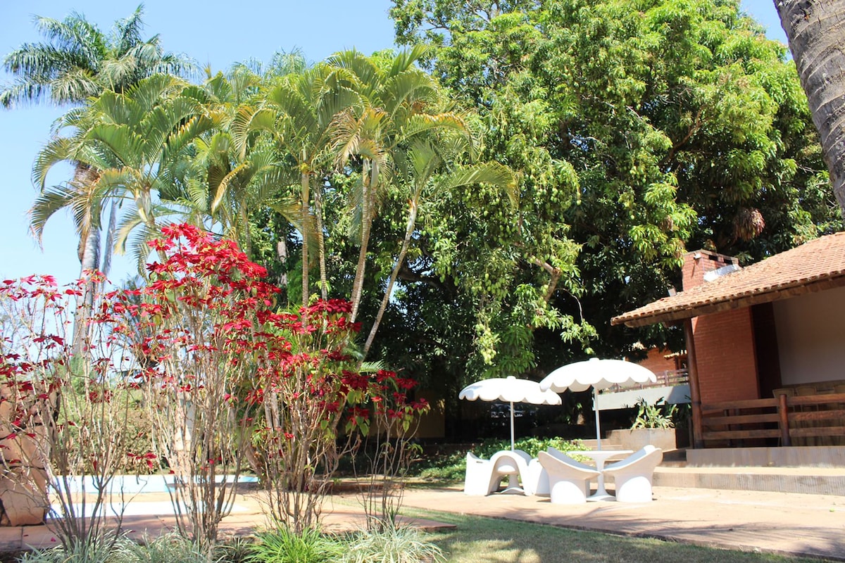 Lush greenery surrounds a spacious outdoor area featuring vibrant red flowers and tall palm trees. A shaded seating area with white umbrellas rests on a paved surface next to a well-maintained lawn, inviting relaxation in a serene environment.