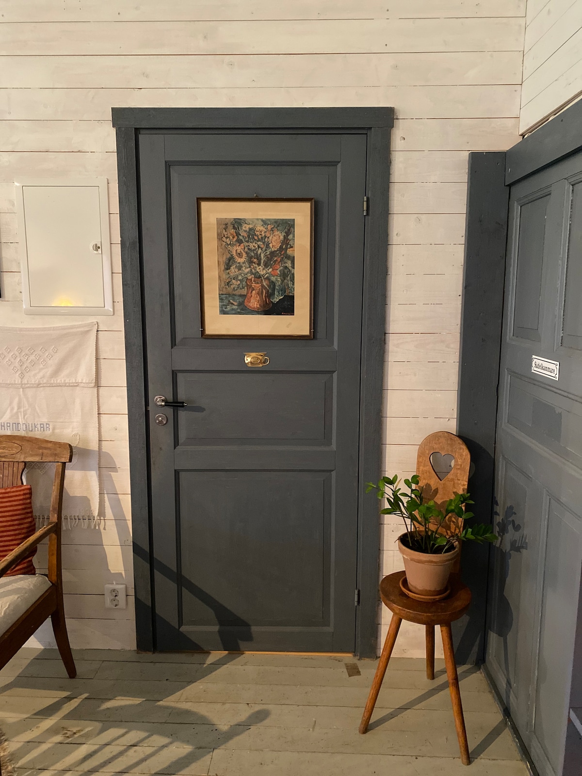A gray door with a brass handle is featured prominently on a white wooden wall. Above the door, a framed floral artwork enhances the space. A small potted plant sits on a wooden chair beside a circular stool, adding a touch of greenery.