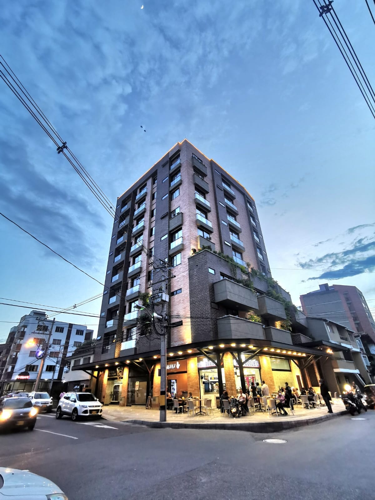 A modern six-story building is positioned at a street corner, featuring a mix of brick and glass exteriors. Outdoor seating areas and greenery are visible at the entrance, while the sky reflects a deepening blue hue as evening approaches, casting soft light on the surroundings.