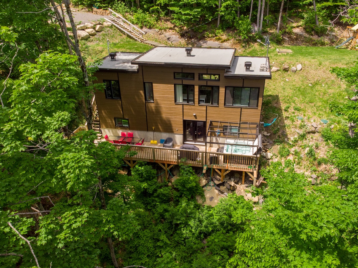 A contemporary chalet sits elevated among lush greenery, featuring large windows for ample natural light. A deck is visible, showcasing a hot tub and seating arrangements. Surrounding trees create a sense of seclusion and connection to nature, complementing the serene landscape.