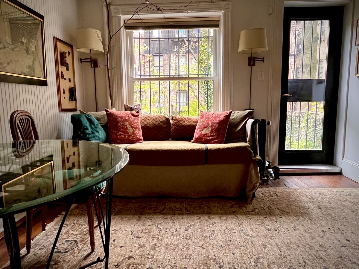 A cozy common room is visible, featuring a comfortable sofa with decorative pillows in front of a large window. Natural light filters in, illuminating the room with a welcoming atmosphere. A glass dining table sits nearby, complementing the wooden floor and soft area rug.