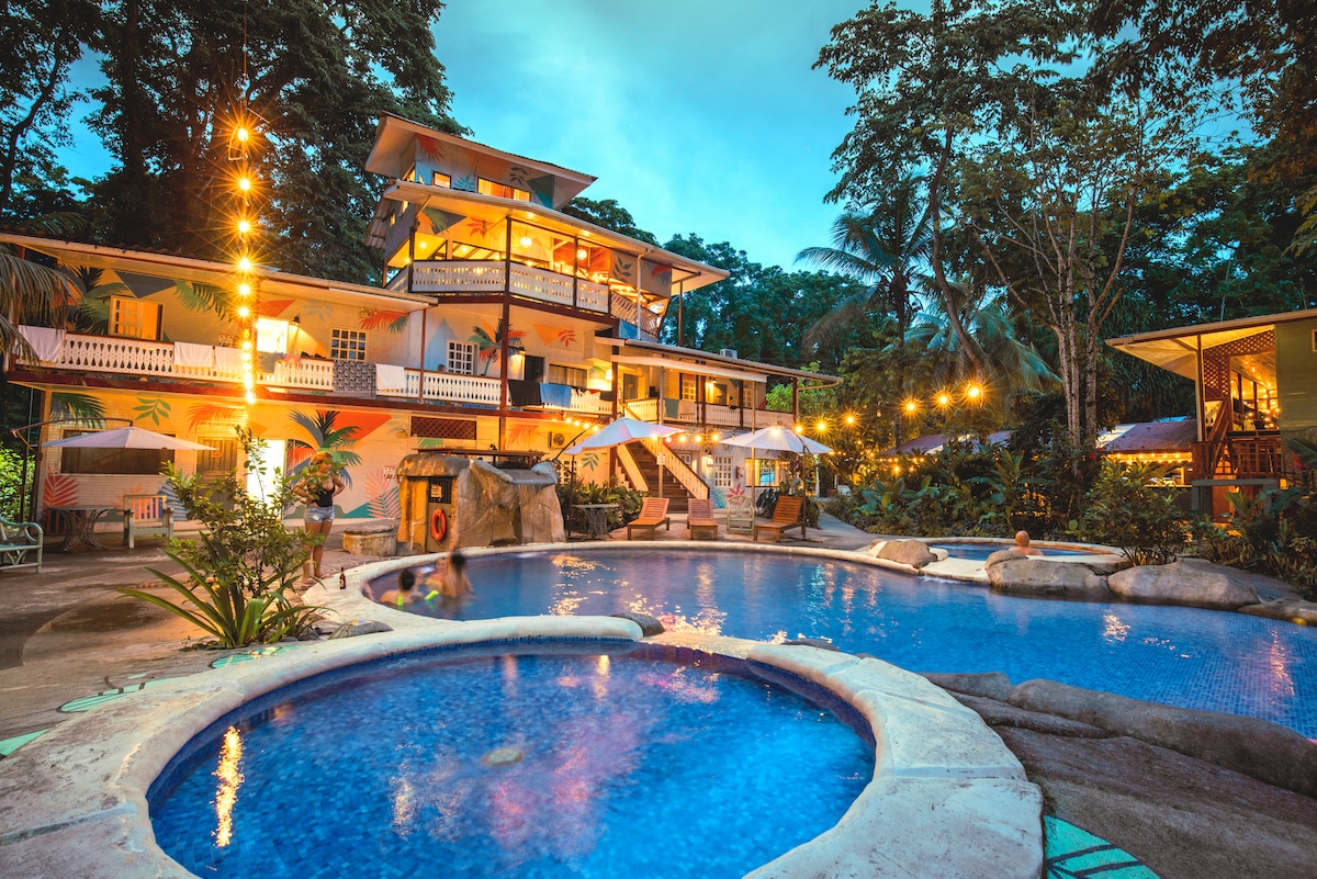 A multi-story building is framed by lush greenery, featuring a large swimming pool at the forefront. Warm lights illuminate the structure and surrounding areas, enhancing the inviting atmosphere. A jacuzzi is positioned beside the pool, accompanied by lounge chairs and umbrellas for relaxation.