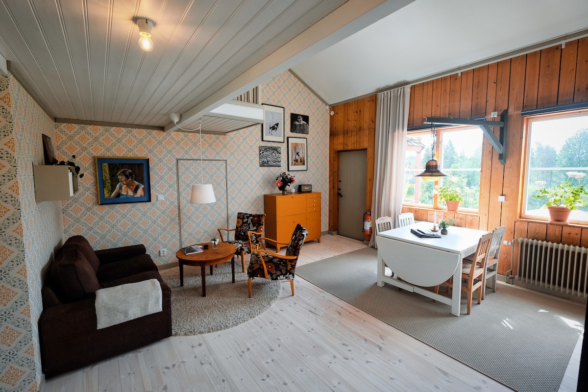 A cozy living space features a brown sofa paired with an armchair in a floral pattern. Natural light floods in through large windows adorned with plants. A dining table for six and a sideboard enhance the sense of comfort, while patterned wallpaper adds a touch of character.
