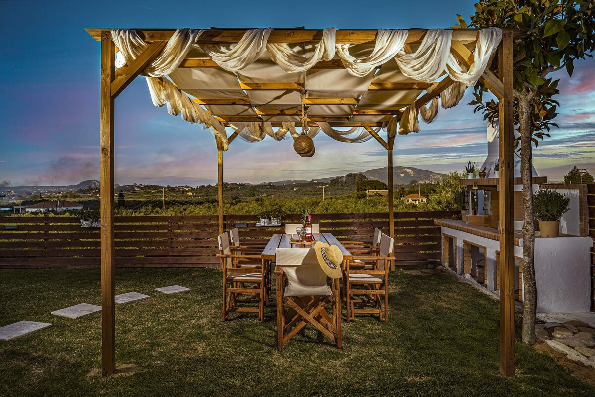 An outdoor dining area features a wooden pergola draped with soft fabric, creating a shaded space. A long table is set with chairs, surrounded by a lush green lawn. Scenic countryside views are visible in the background, complemented by a gentle sunset.
