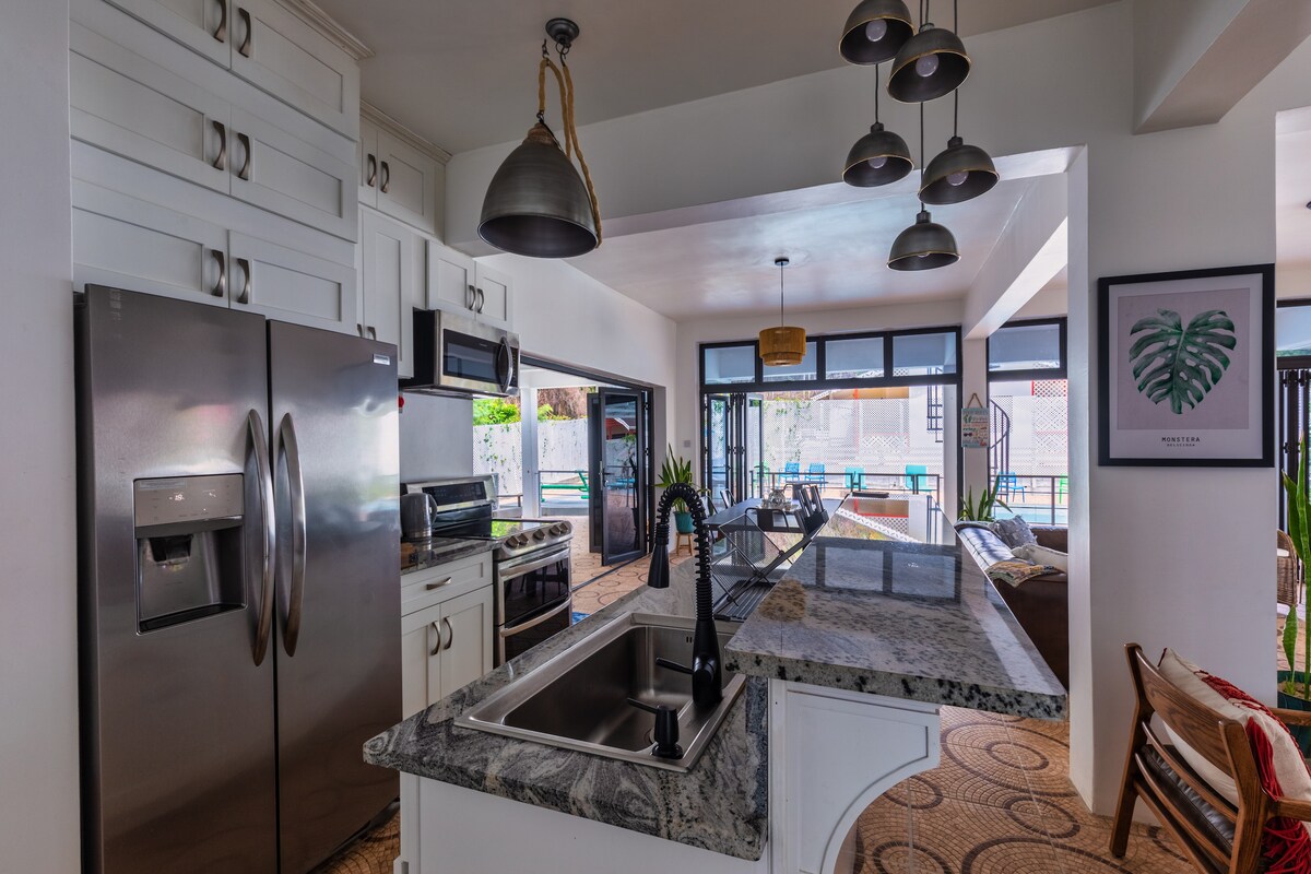 A modern kitchen features stainless steel appliances, including a large refrigerator and a gas stove. Granite countertops enhance the workspace, while pendant lights hang above the island sink. Bifold doors open to connect the kitchen with the outdoor pool area, creating a seamless flow between spaces.
