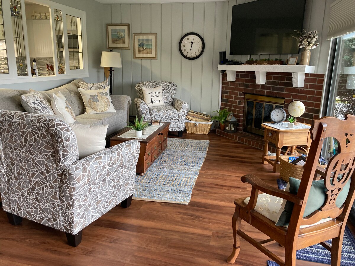 A comfortable living area is presented with two armchairs and a sofa, arranged around a wooden coffee table. A flat-screen TV is mounted above a brick fireplace. Decorative accents, including a wall clock and art pieces, contribute to a cozy ambiance.