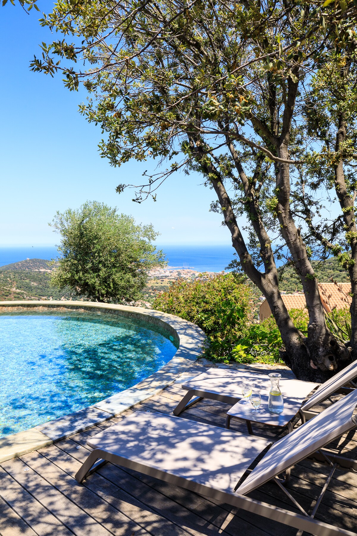 A circular pool is surrounded by a stone deck and shaded by trees. Two lounge chairs are positioned beside the water, offering a view of the distant coastline and the calming blue sea, complemented by a lush landscape.