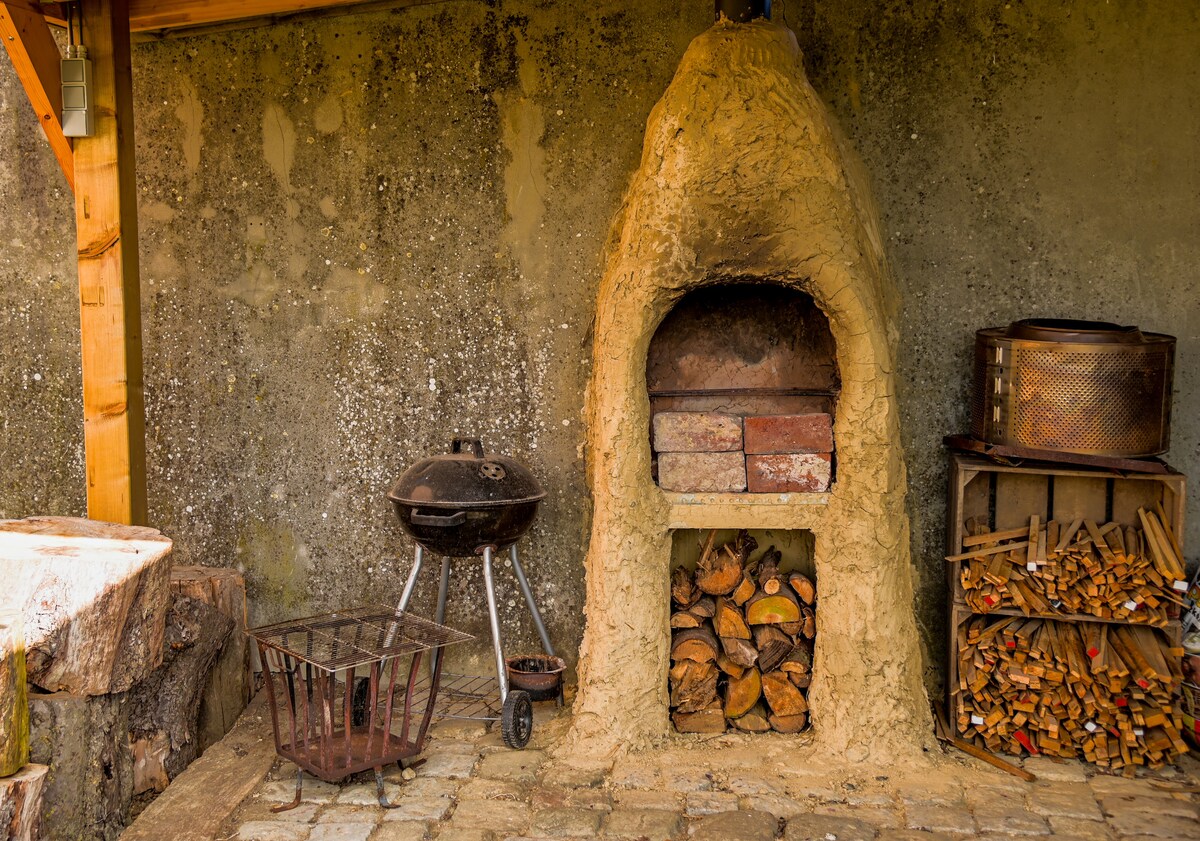 An outdoor cooking area features a traditional stone pizza oven built into a textured wall. A charcoal grill stands next to a stack of firewood, while a rustic wood basket occupies the space. The stone-paved floor complements the natural surroundings.
