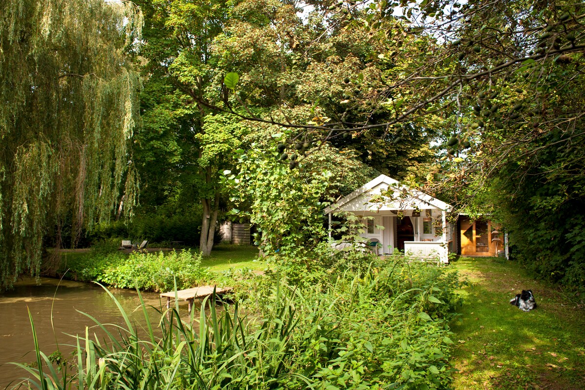 A serene view of two charming cottages nestled among lush greenery alongside a tranquil pond. The area is framed by trees and plants, providing a sense of privacy. A small wooden platform extends towards the water, creating a peaceful outdoor space.