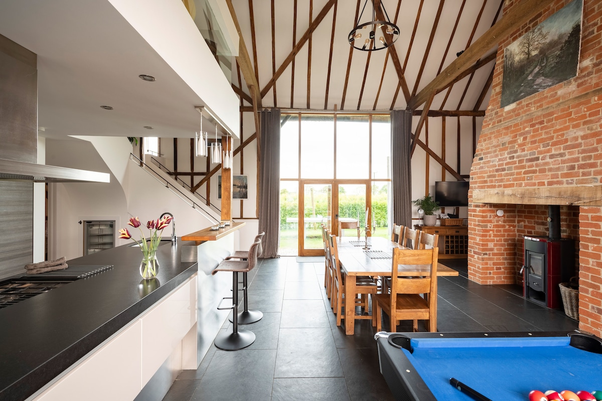An open-plan living area showcases high wooden beams and large windows, allowing natural light to fill the space. A dining table for eight is visible alongside a comfortable seating arrangement. A pool table is elegantly placed in one corner, complemented by a fully equipped kitchen.