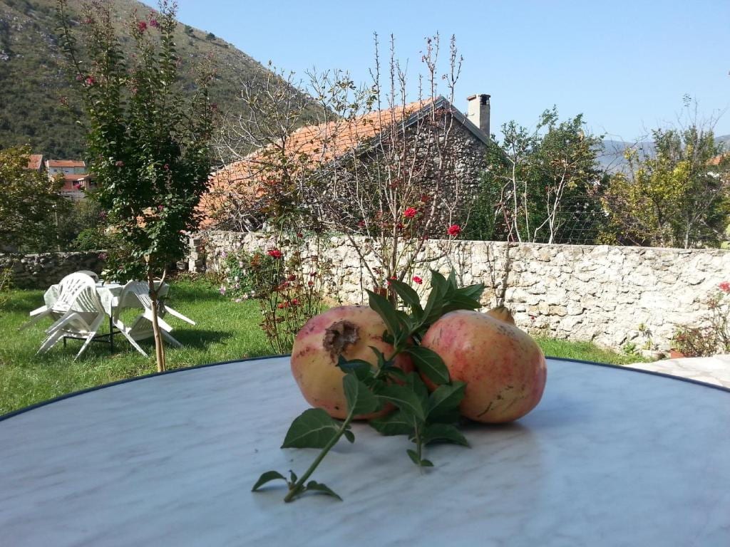 Two fresh pomegranates are placed on a marble table, surrounded by a lush green garden. In the background, charming stone houses and blooming flowers are visible under a clear blue sky. Chairs are positioned nearby, offering a serene outdoor space.