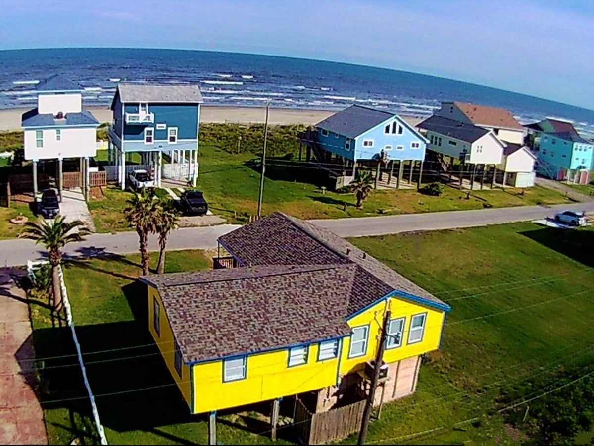An aerial view captures The Skipper with its bright yellow exterior, surrounded by green grass and palm trees. The ocean waves can be seen beyond a quiet street lined with colorful beach homes, emphasizing the proximity to the beach.